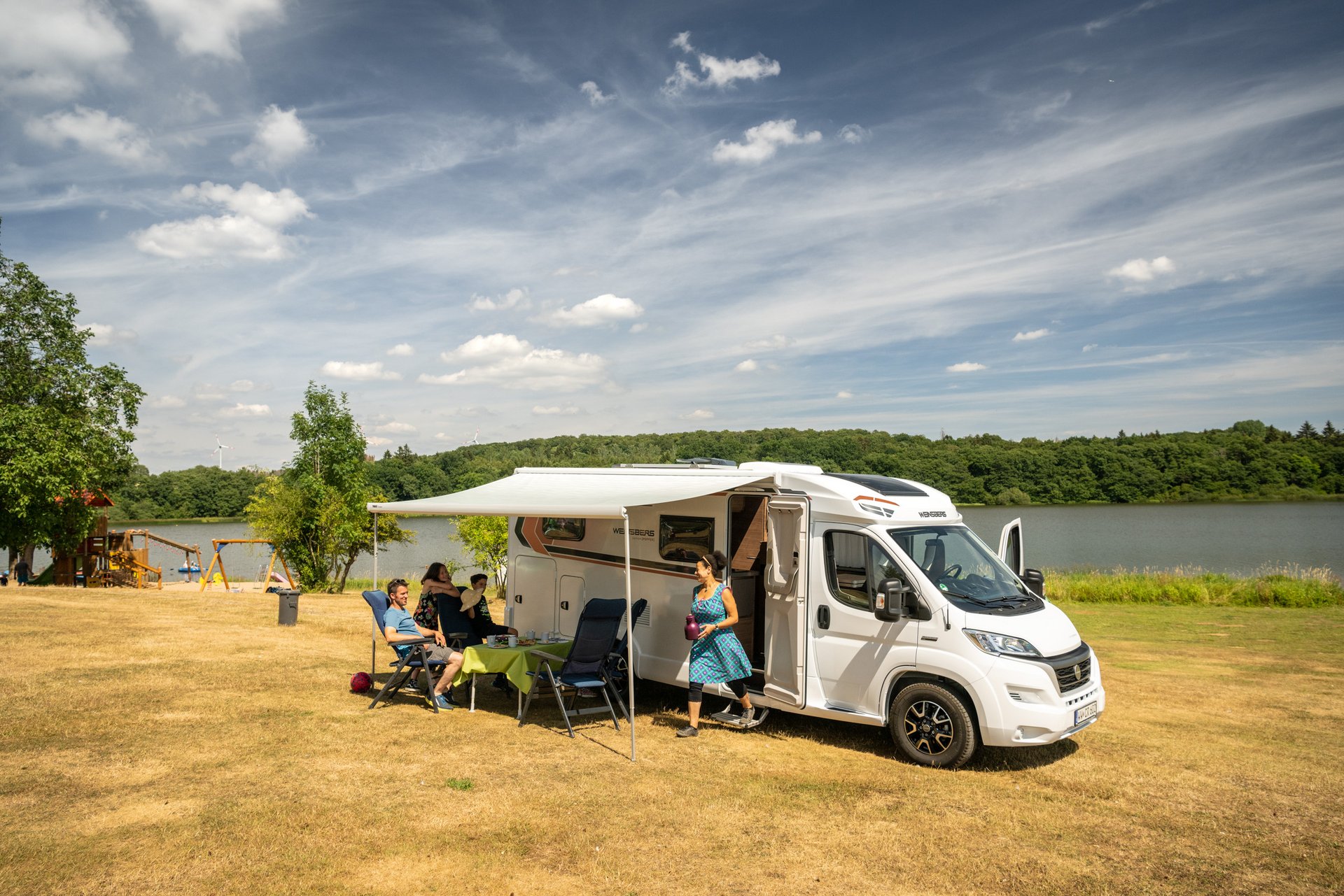 Familie entspannt vor ihrem Wohnmobil am ruhigen See bei sonnigem Wetter